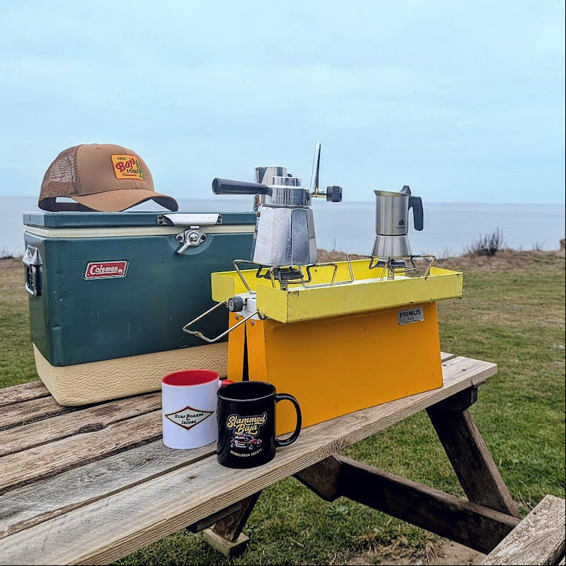 Campsite setup with Coleman cooler, coffee maker, and Jacobs Surfboards / Slammed Baja Appreciation Society mugs on a wooden table outdoors. Caramel 1973 Baja 1000 cap on top of Coleman cooler.
