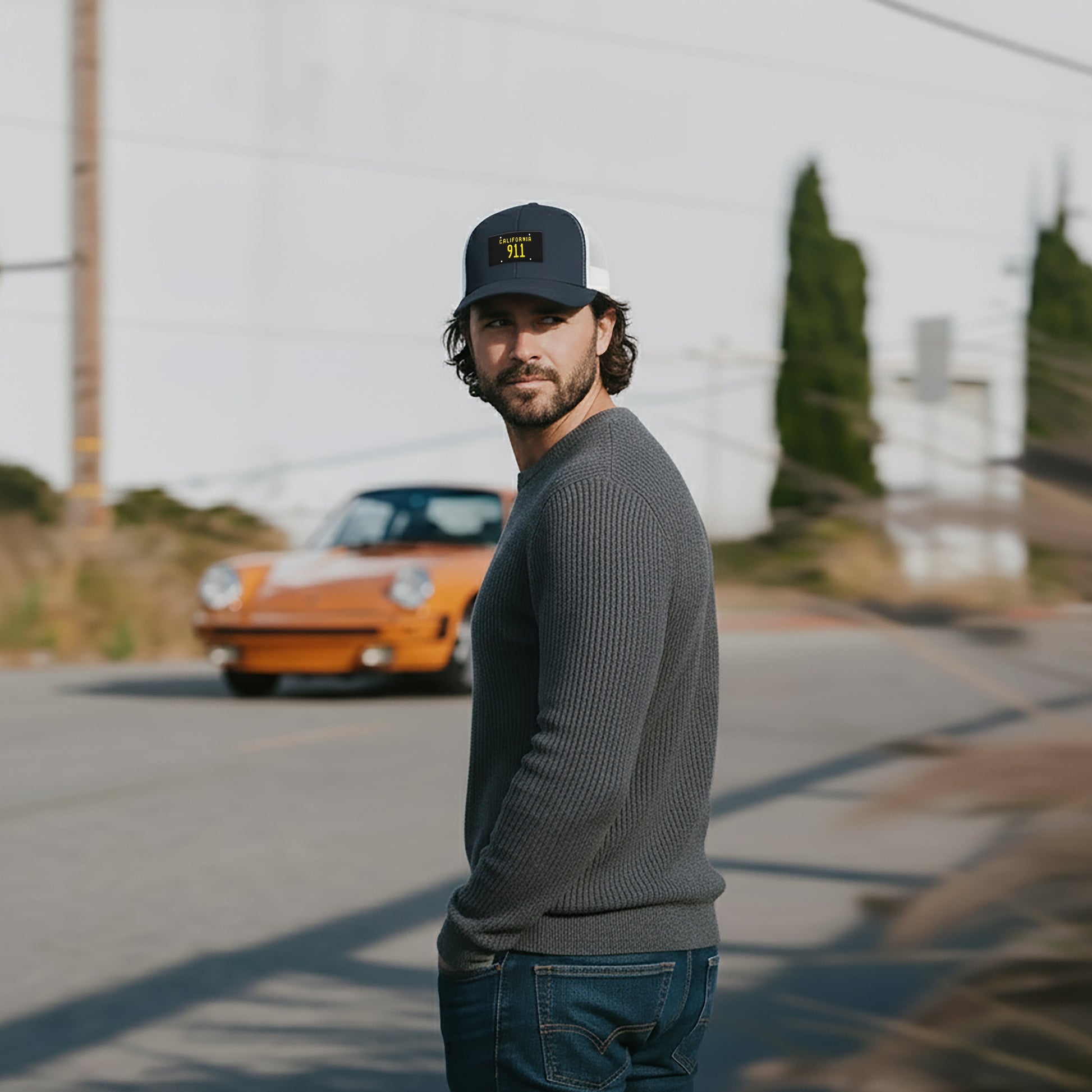 Man wearing a California Black plate 911 Trucker cap and sweater standing on a road with an orange Porsche 911 in the background
