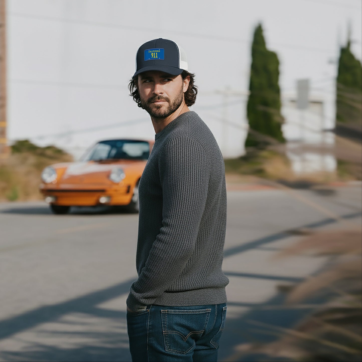 Man wearing a California 911 Trucker Hat standing on a road with an orange Porsche 911 in the background.