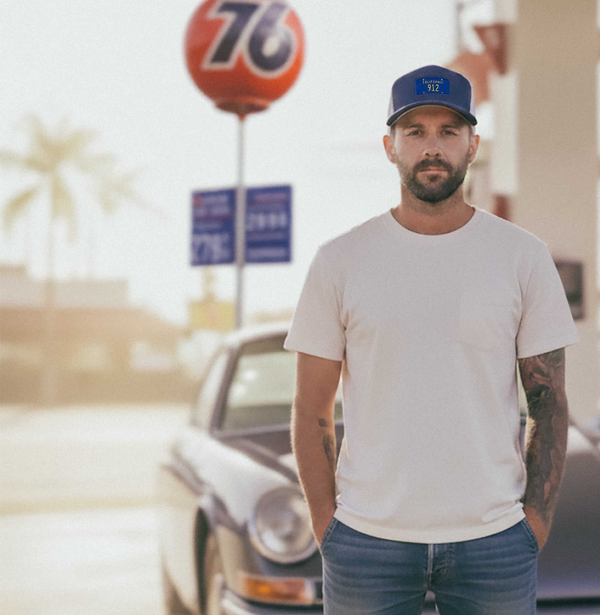 Man standing in front of a vintage Porsche at a Union 76 gas station, wearing a California 912 Trucker hat. 