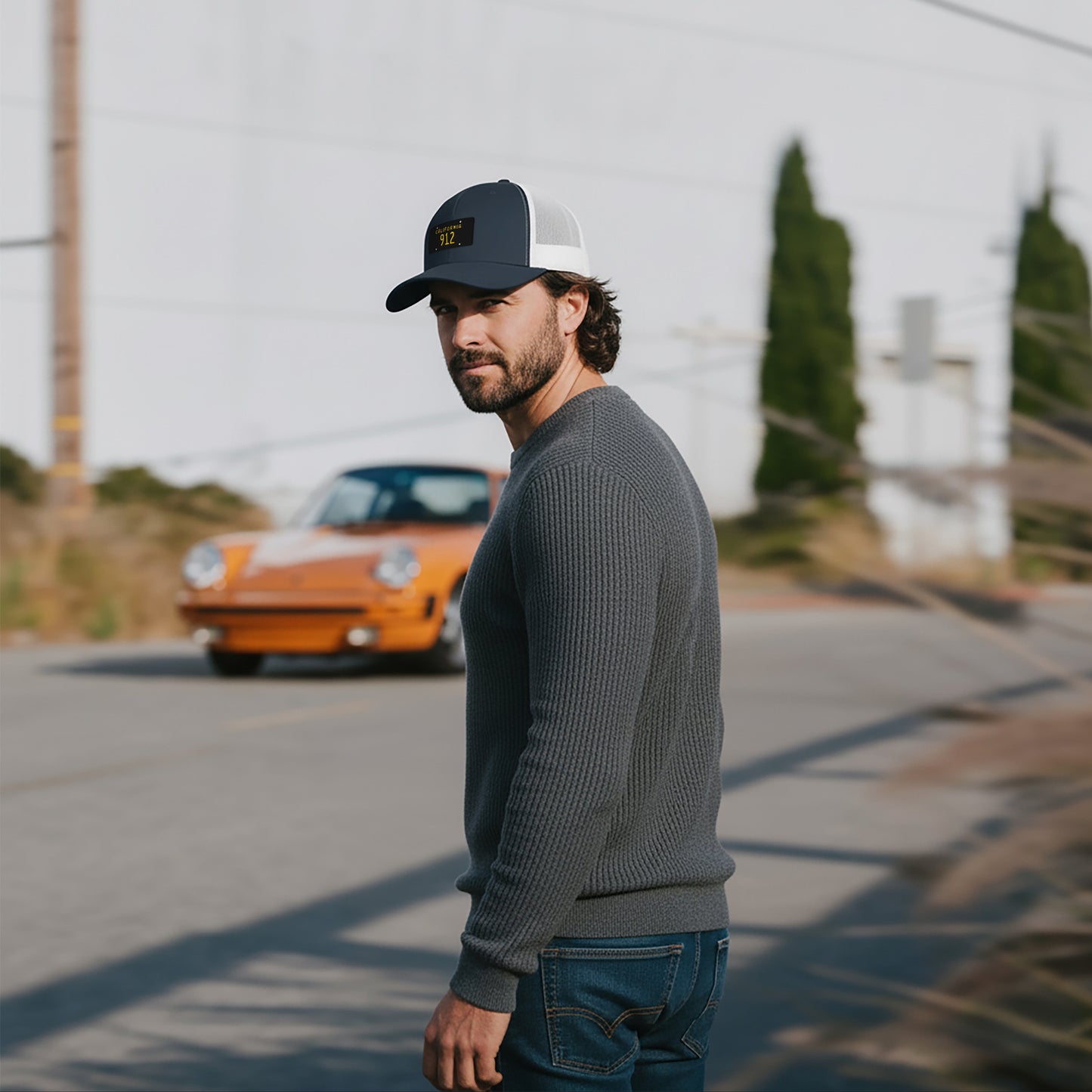 Man wearing a California black plate 912 Trucker hat and sweater standing on a road with a vintage orange Porsche 911 in the background.