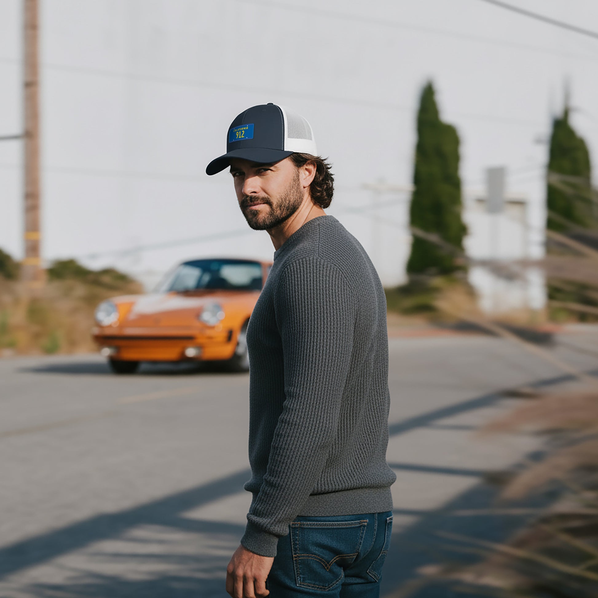 Man wearing a Porsche 912 cap and gray sweater standing on a road with an orange Porsche 911 in the background.