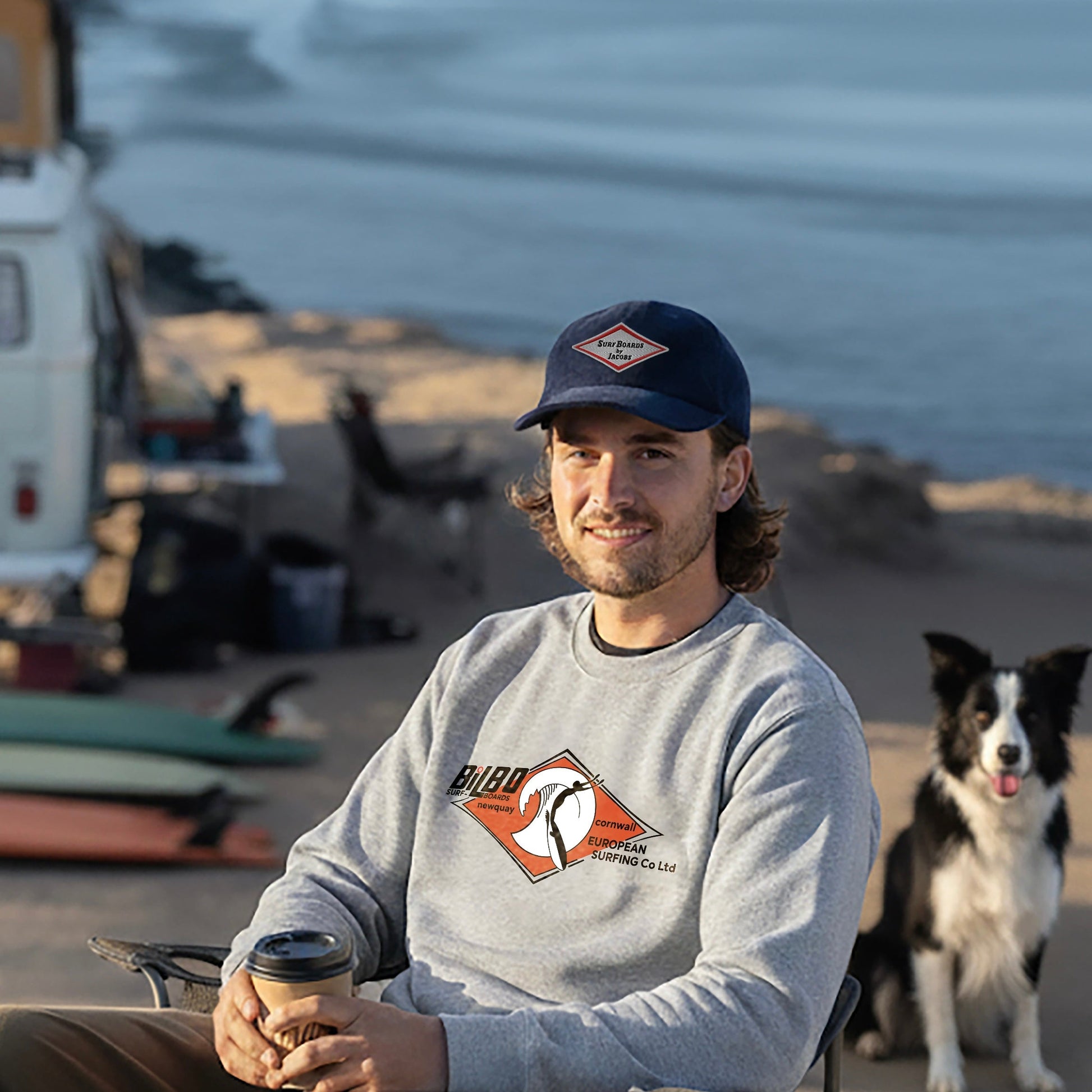 Man sitting on a beach with a dog, VW Bus and Surfboards, holding a coffee cup, wearing a Bilbo Surfboards sweatshirt and Surfboards by Jacobs Corduroy cap in Oxford Blue.