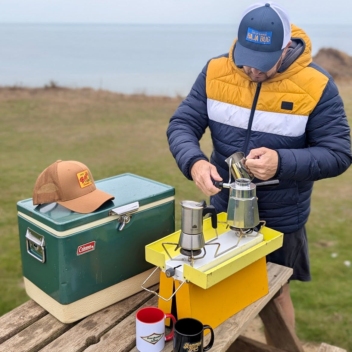 Person preparing coffee outdoors with a Coleman cooler and camping stove. Man wears the Navy/white California Baja Bug trucker hat. The Caramel 1973 Baja 1000 trucker hat sits atop the cooler. The Jacobs Surfboards mug and Slammed Baja Mug are waiting to be filled with coffee.