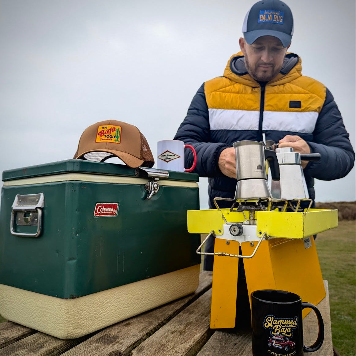 Man preparing coffee using a portable stove with a Coleman cooler. Man wears the California Baja Bug Trucker Hat, with the Caramel 1973 Baja 1000 trucker hat and Slammed Baja Appreciation Society mug in the image also.
