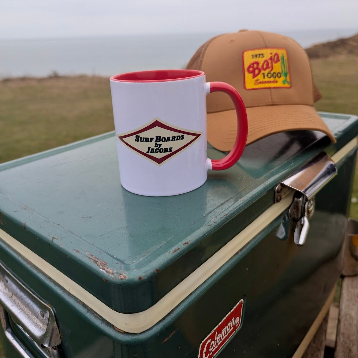 White/red Surfboards by Jacobs mug with red handle on a vintage green Coleman cooler outdoors, with a Caramel 1973 Baja 1000 Trucker Hat.