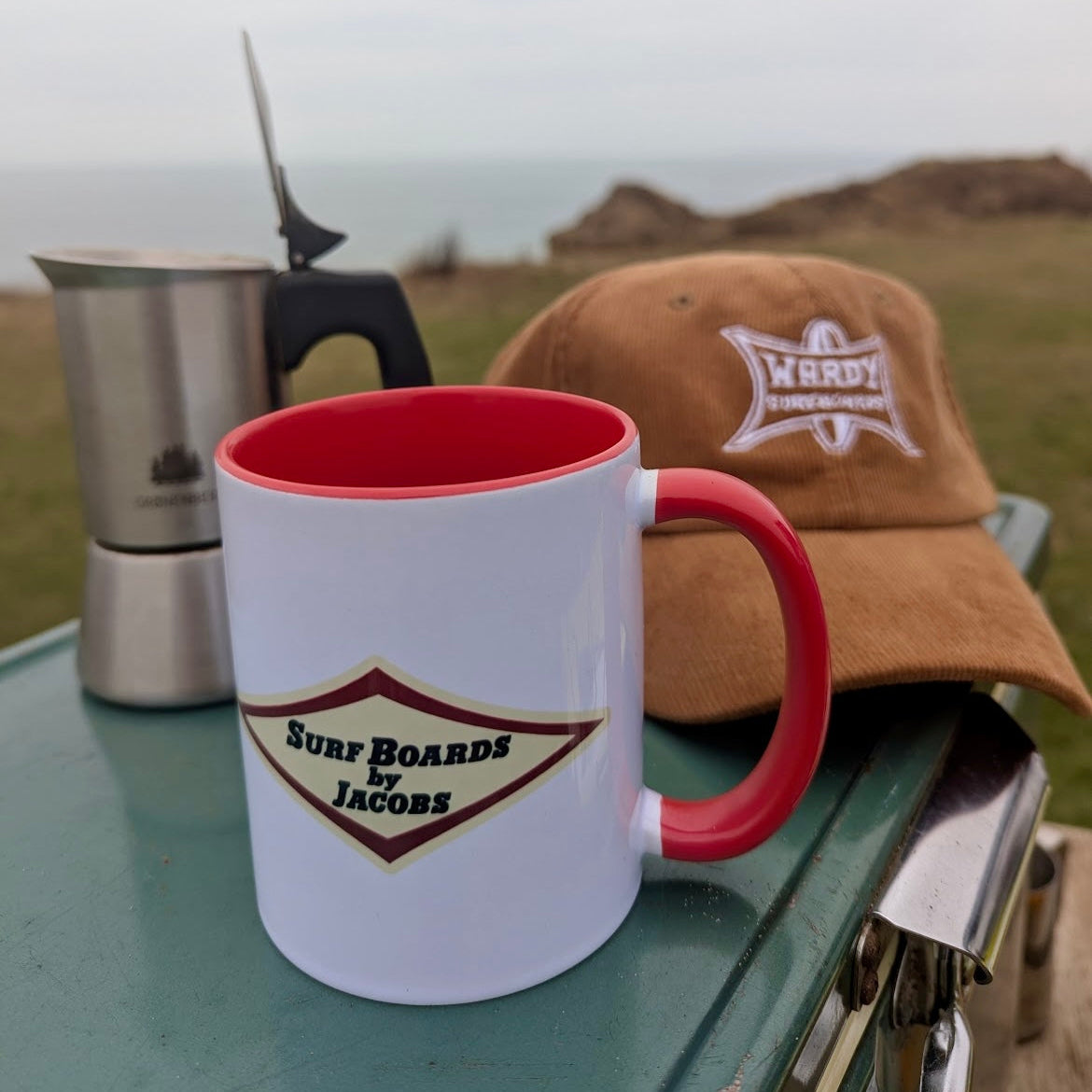 White Surf Boards by Jacobs mug with red interior and handle, featuring 'Surf Boards by Jacobs' logo, on a table outdoors. Wardy Surfboards cord cap in the background.