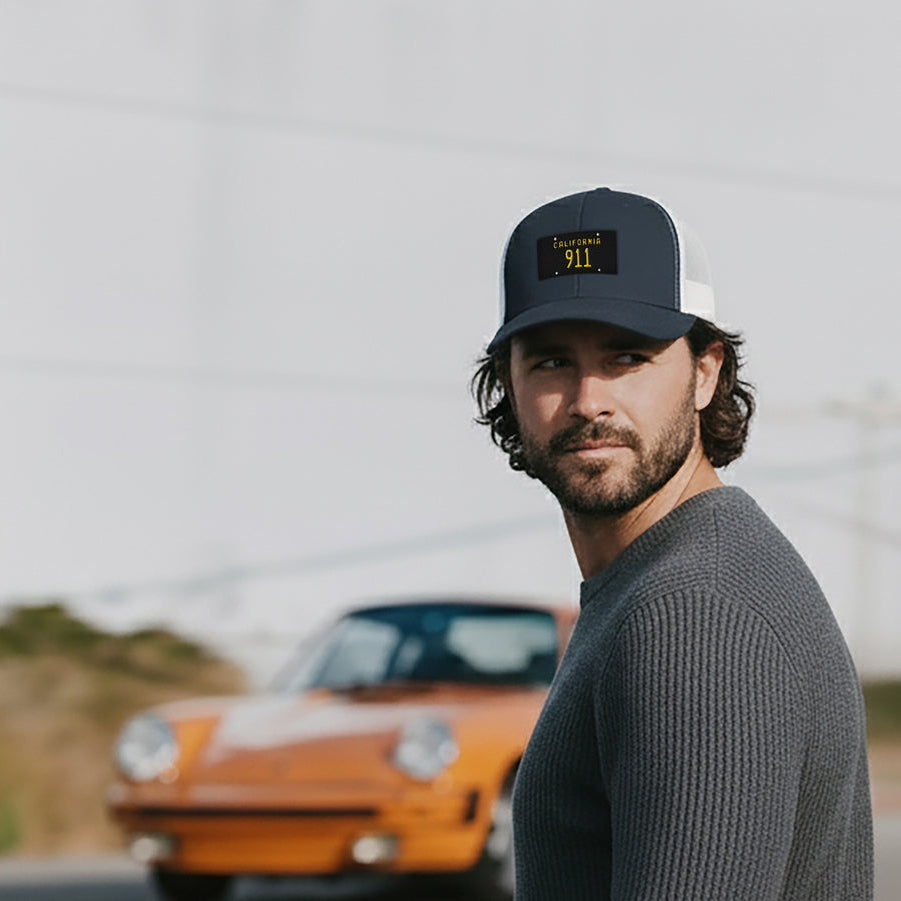 Man wearing a cap with the California black plate 911 embroidered logo in front of an orange Porsche 911.