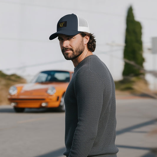 Man wearing a California black plate 912 Trucker hat and sweater standing on a road with a vintage orange Porsche 911 in the background.