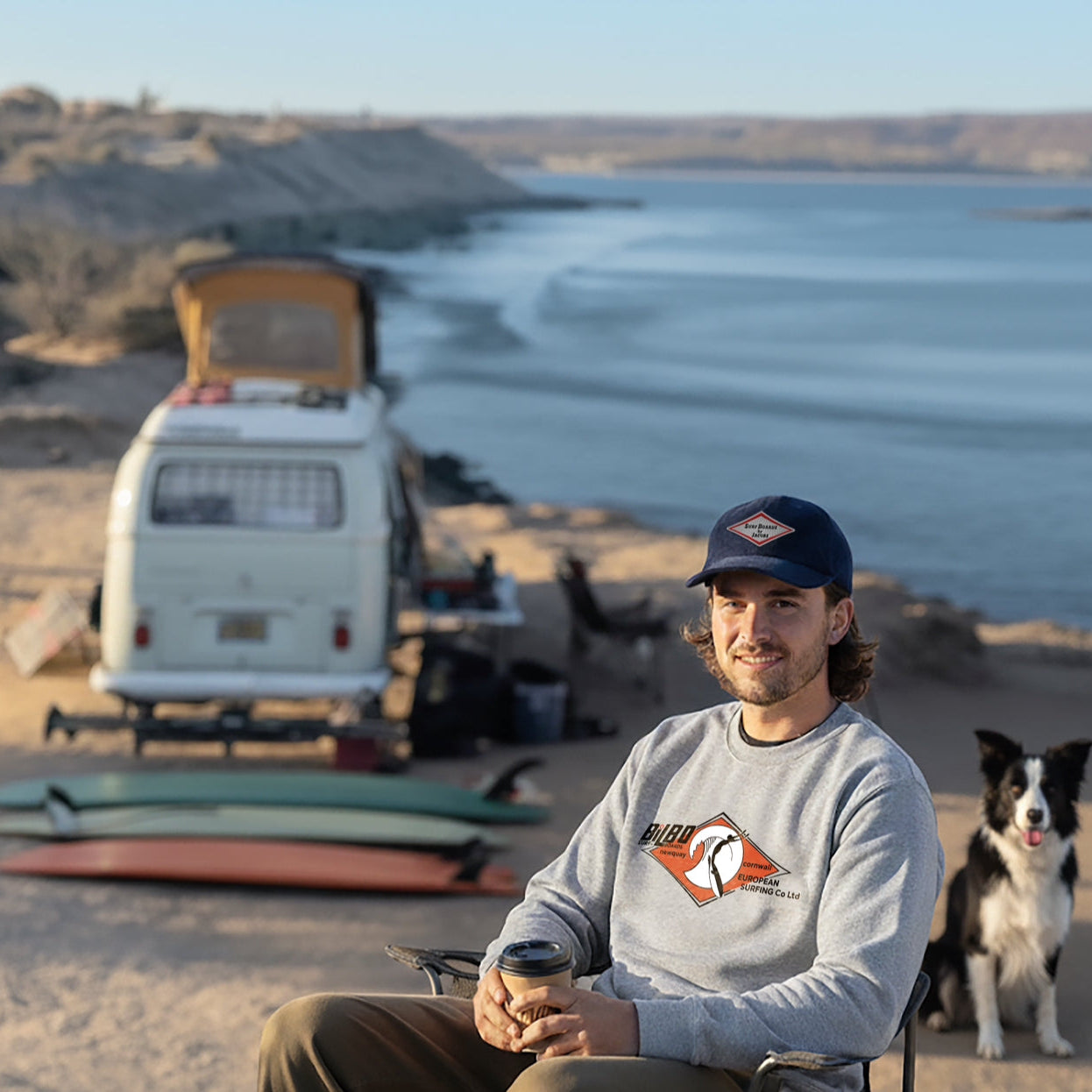 Man sitting on a beach with a dog and a VW Bus in the background. Man wears: Surfboard by Jacobs Oxford Blue Cord Cap and Heather Grey Bilbo Surfboards Sweatshirt.