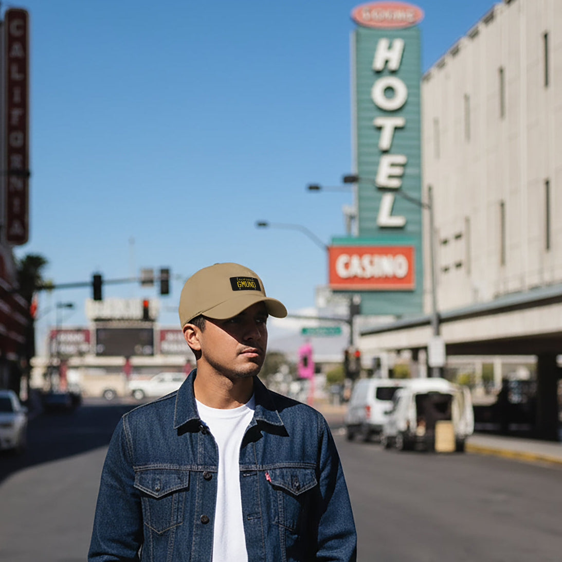 Man wearing a California GMUND Porsche Baseball cap and denim jacket standing on a street with a hotel sign in the background.