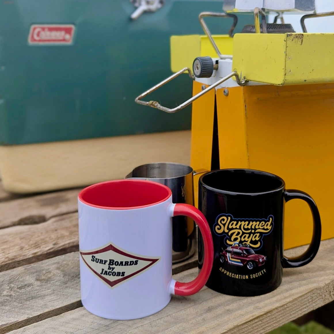 White Surf Boards by Jacobs mug with red interior and handle, featuring 'Surf Boards by Jacobs' logo, on a table outdoors. Slammed Baja Appreciation Society black mug sits next to it.