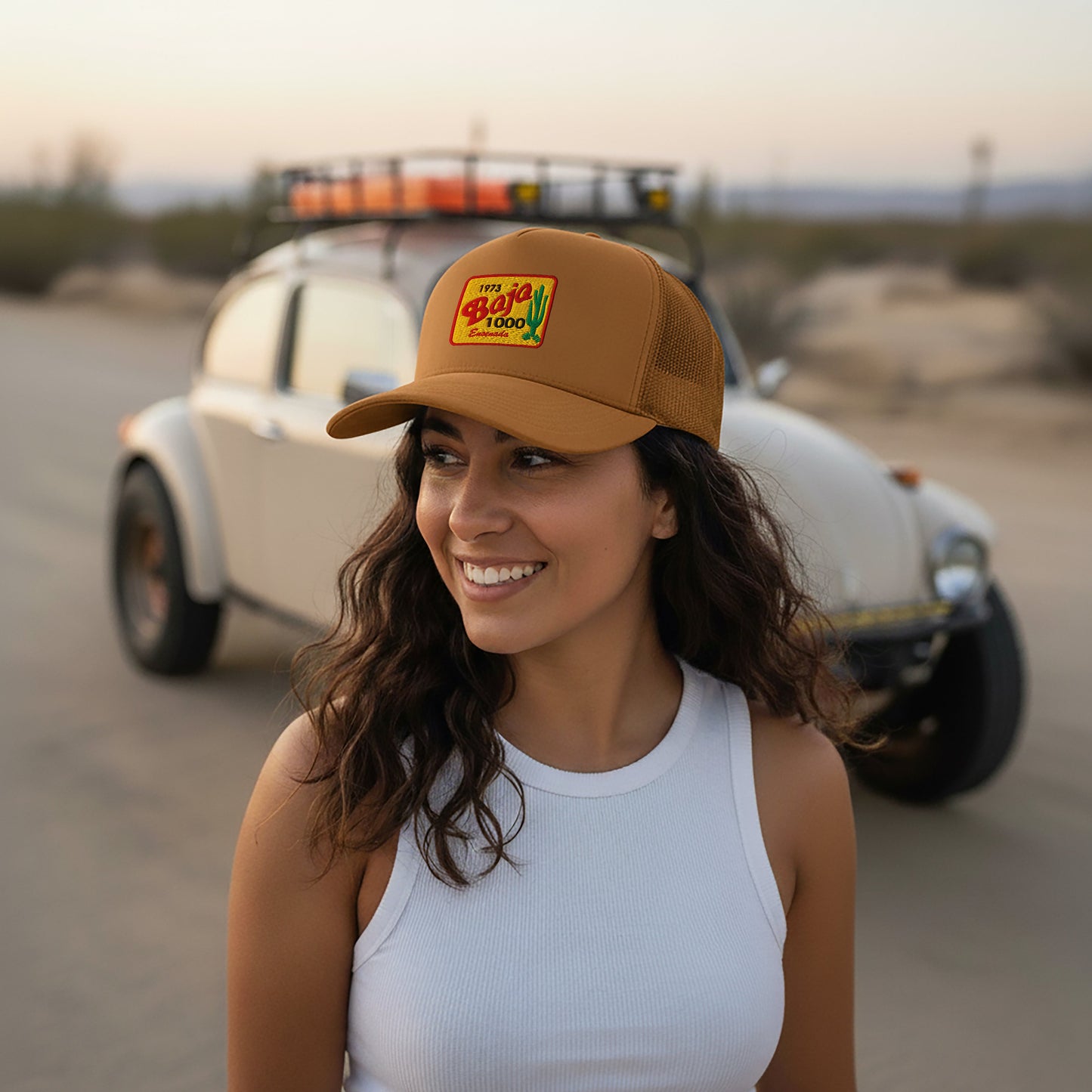 Woman wearing a Caramel 1973 Baja 1000 Trucker Hat standing in front of a vintage Baja Bug in a desert setting