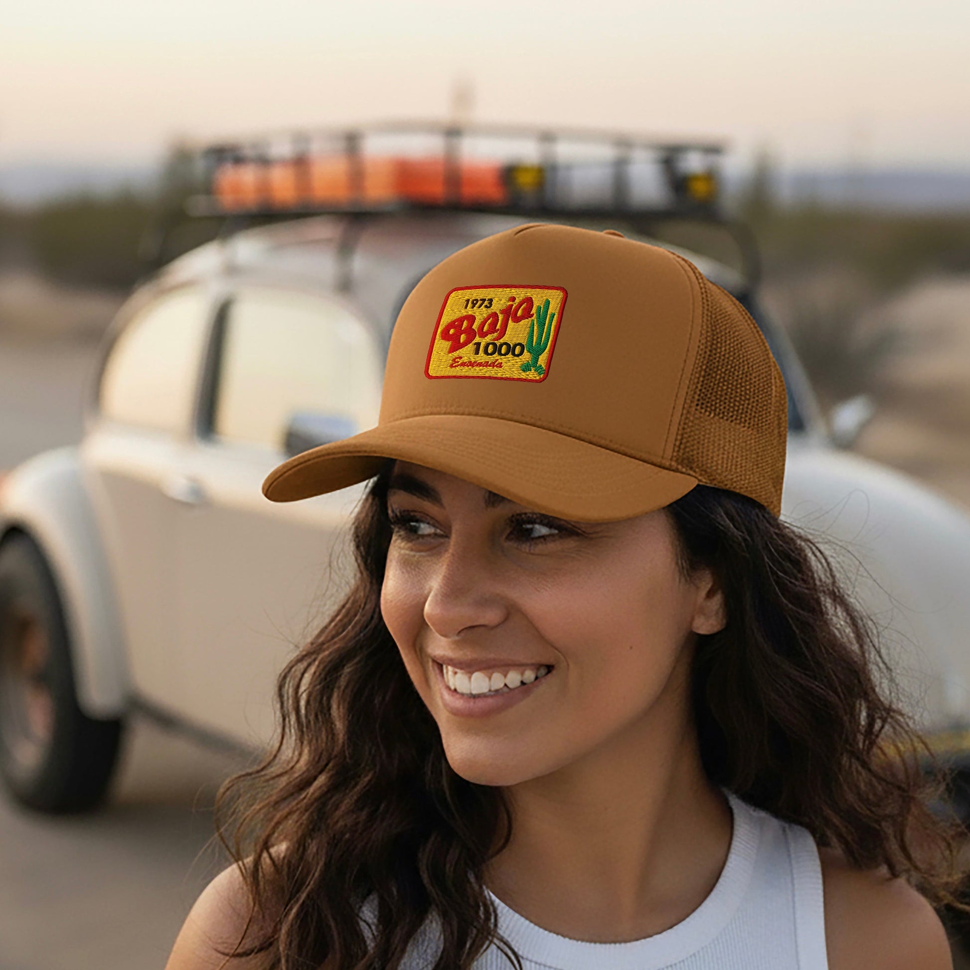 Woman wearing a Caramel 1973 Baja 1000 Trucker Hat, standing in front of a vintage Baja Bug.