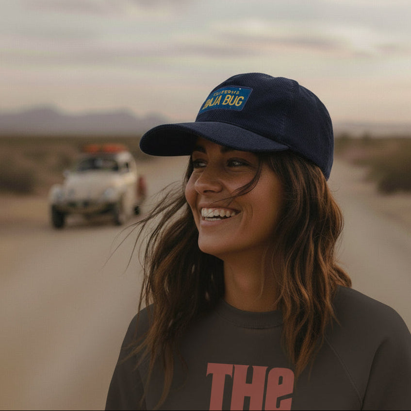 Woman wearing an Oxford Blue corduroy cap with a California BAJA BUG embroidered logo, standing in a desert with a Baja Bug in the background.