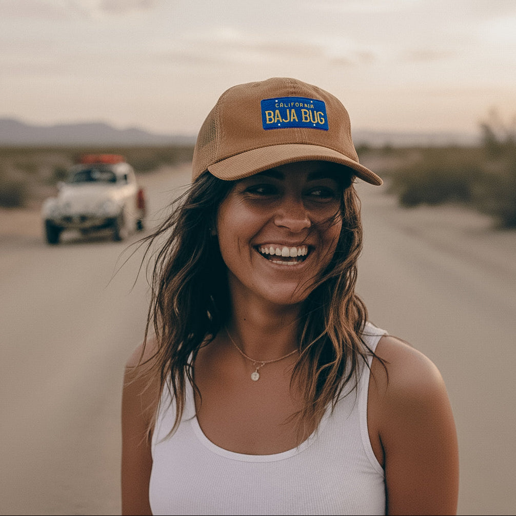 Woman wearing a camel corduroy cap with 'Baja Bug' logo on a desert road with a Baja Bug in the background.