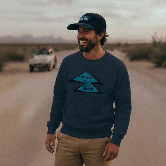 Man wearing a French navy 'Surfboards by The Greek' sweatshirt and an Oxford Blue cord BAJA BUG cap, standing on a desert road with a Baja Bug in the background.