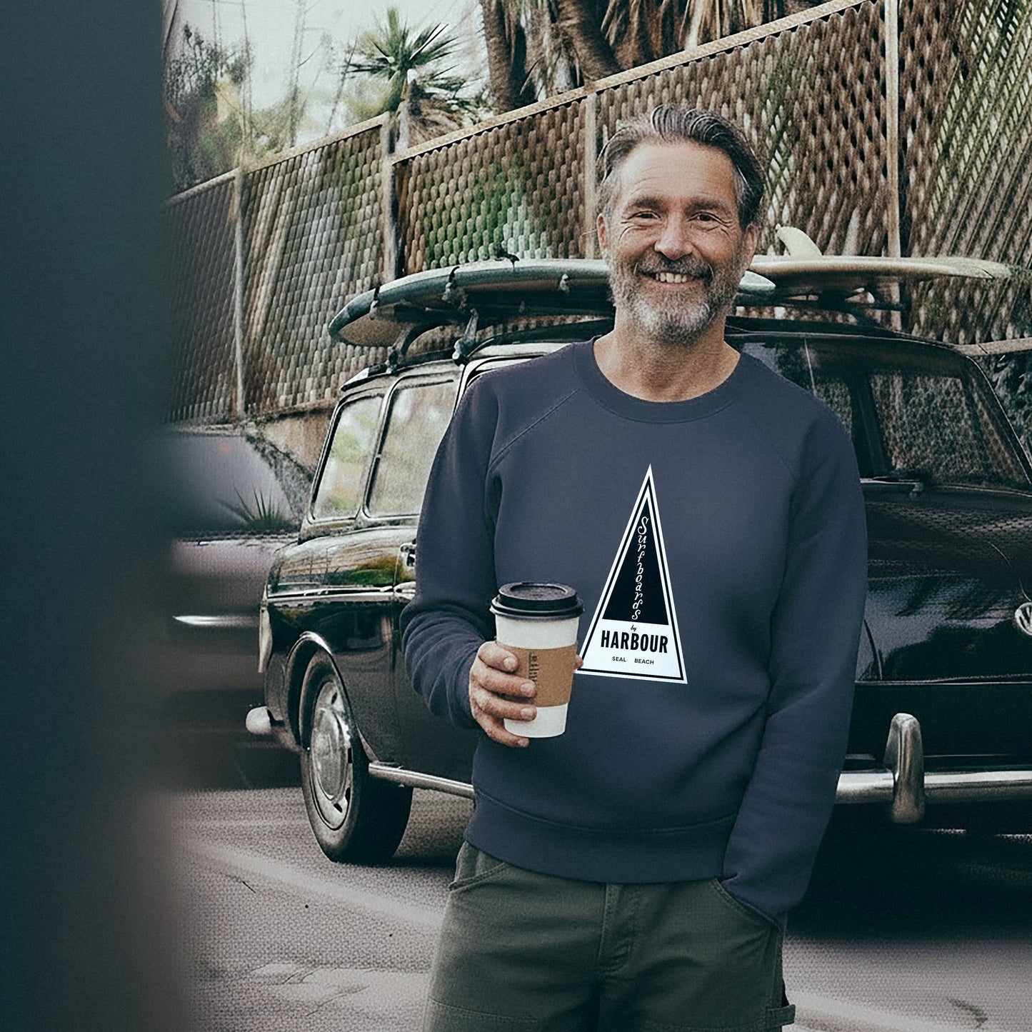 Man wearing a French Blue Harbour Surfboards Sweatshirt holding a coffee cup in front of a VW Squareback with a surfboard on top.