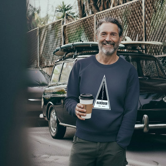 Man wearing a French Blue Harbour Surfboards Sweatshirt holding a coffee cup in front of a VW Squareback with a surfboard on top.