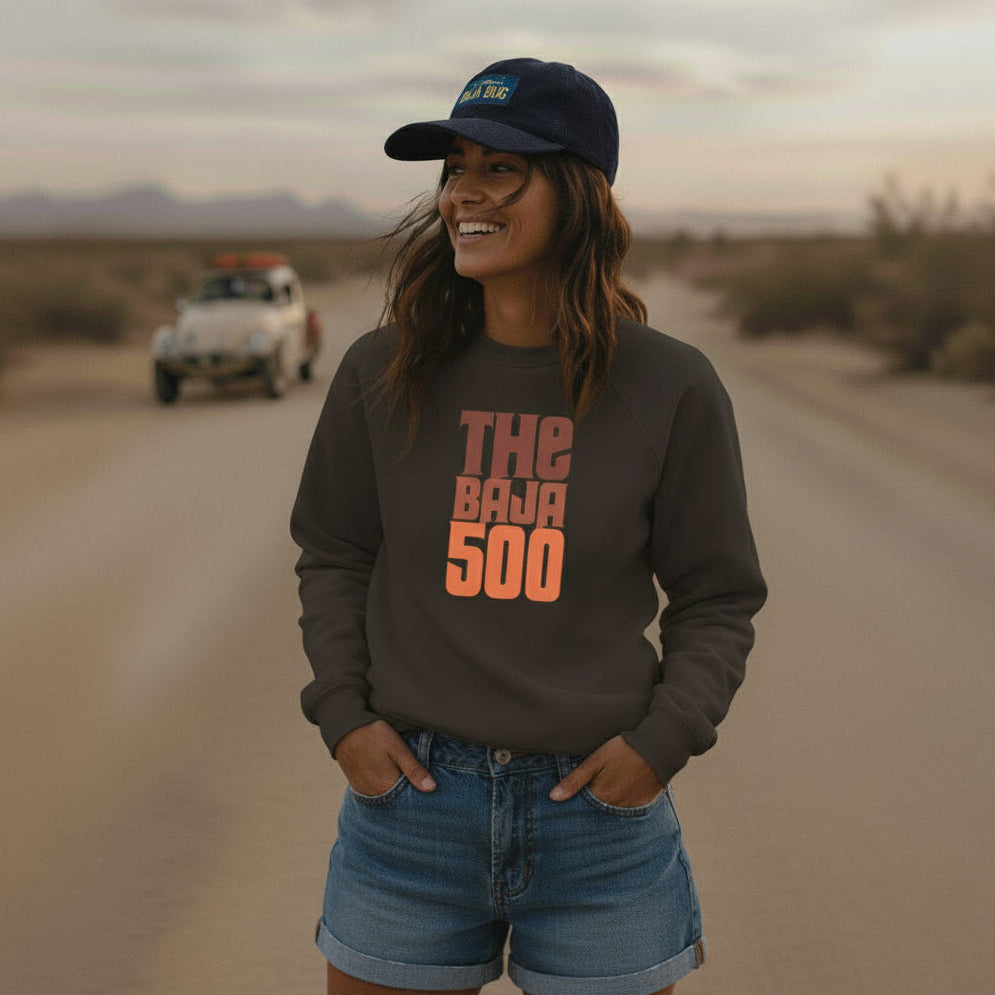 Woman wearing a 'The Baja 500' sweatshirt and an Oxford Blue corduroy California Baja Bug cap on a desert road with a Baja Bug in the background.