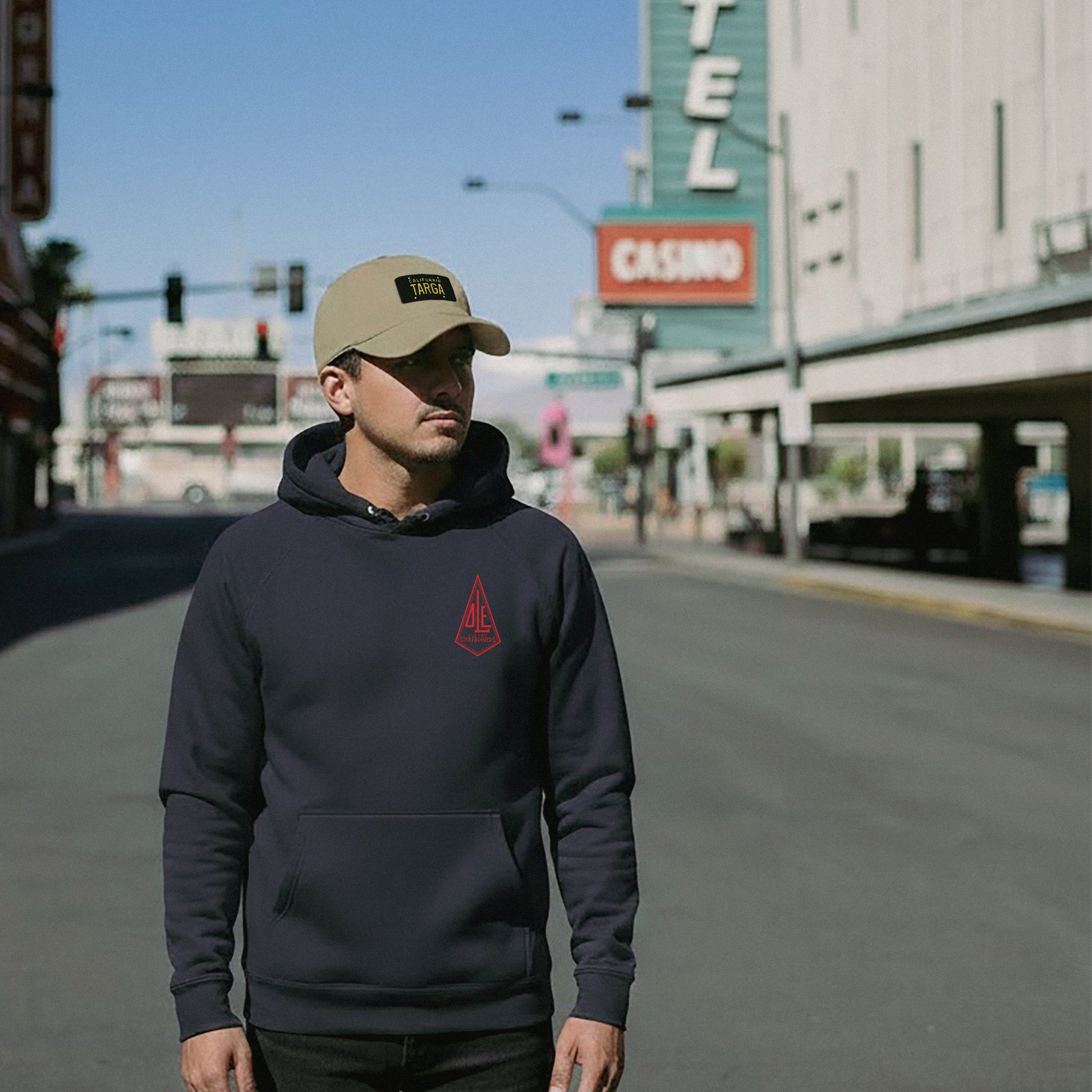 Man wearing a navy Ole Surfboards hoodie and beige California Porsche Targa cap on a city street.