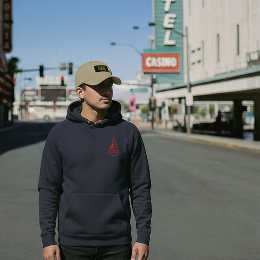 Man wearing a navy Ole Surfboards hoodie and beige California Porsche Targa cap on a city street.
