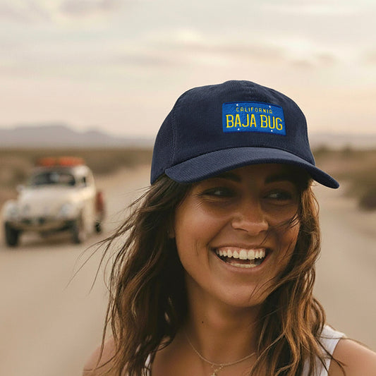 Woman wearing an Oxford blue California Baja Bug corduroy cap with 'Baja Bug' logo in a desert setting with vintage Baja Bug.