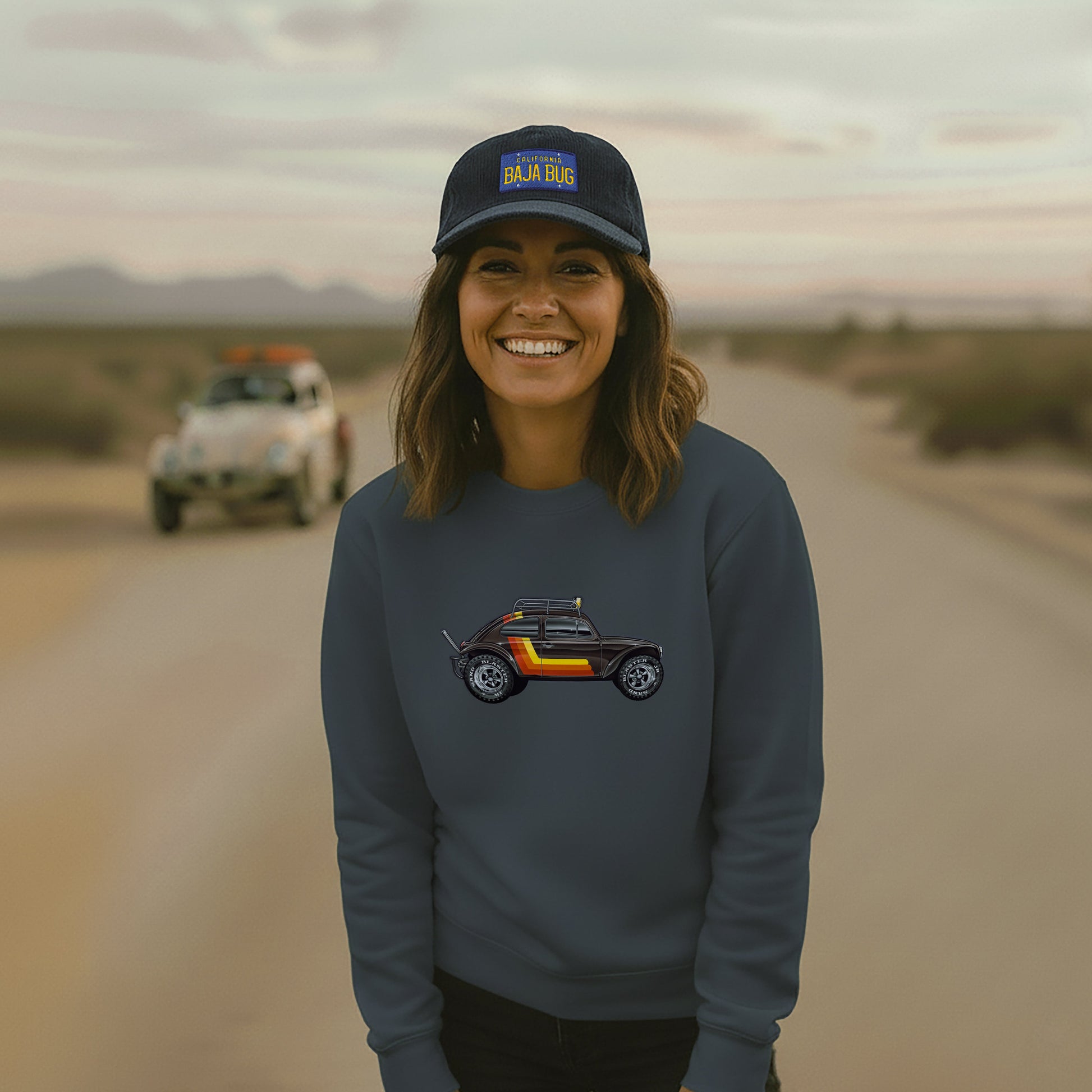 Person wearing a Stripes Baja Bug sweatshirt on a desert road with a Baja Bug in the background.