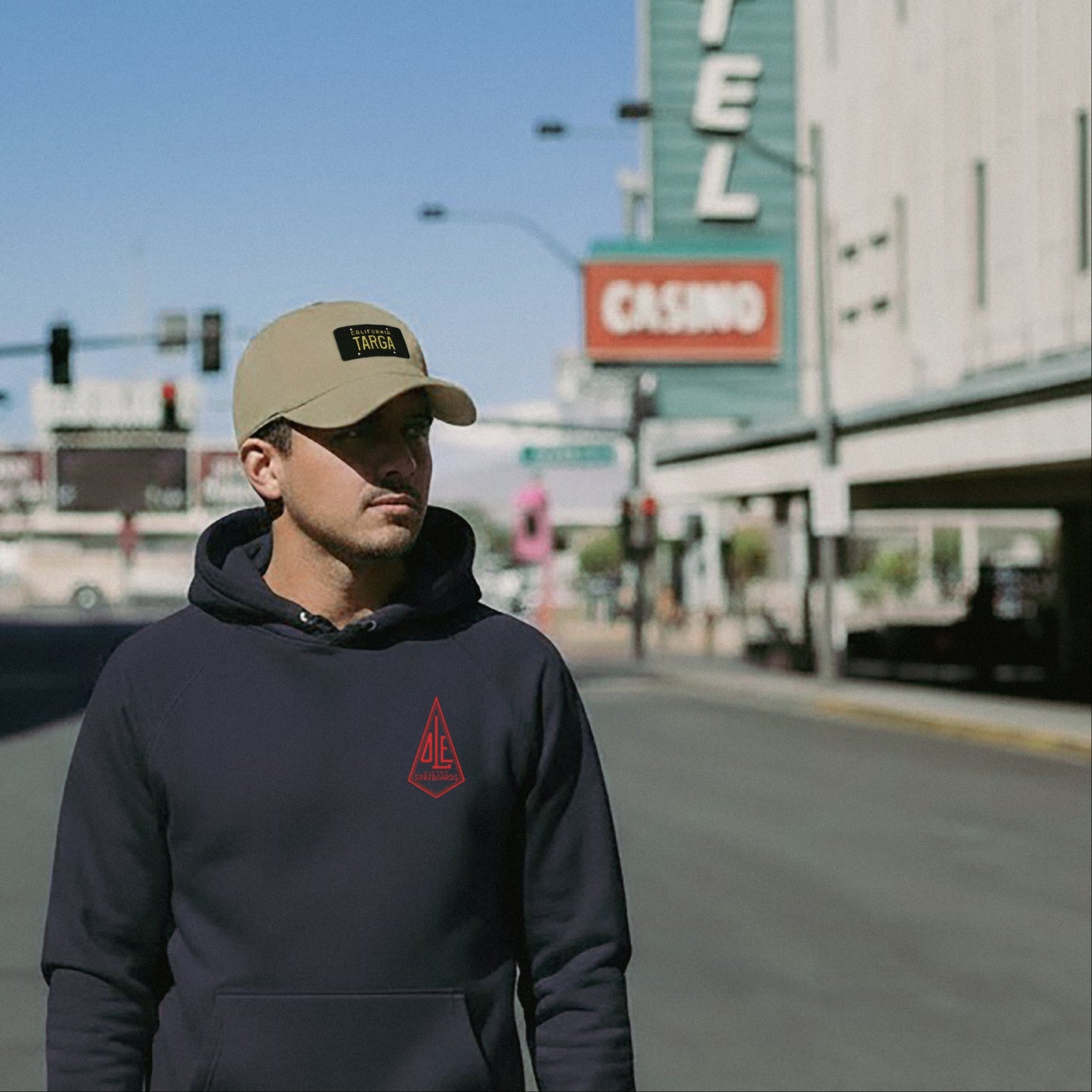 Man wearing a navy Ole Surfboards hoodie and beige California Targa Baseball cap on a city street.