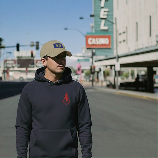 Man wearing a navy Ole Surfboards hoodie and beige California Porsche TARGA cap on a city street.