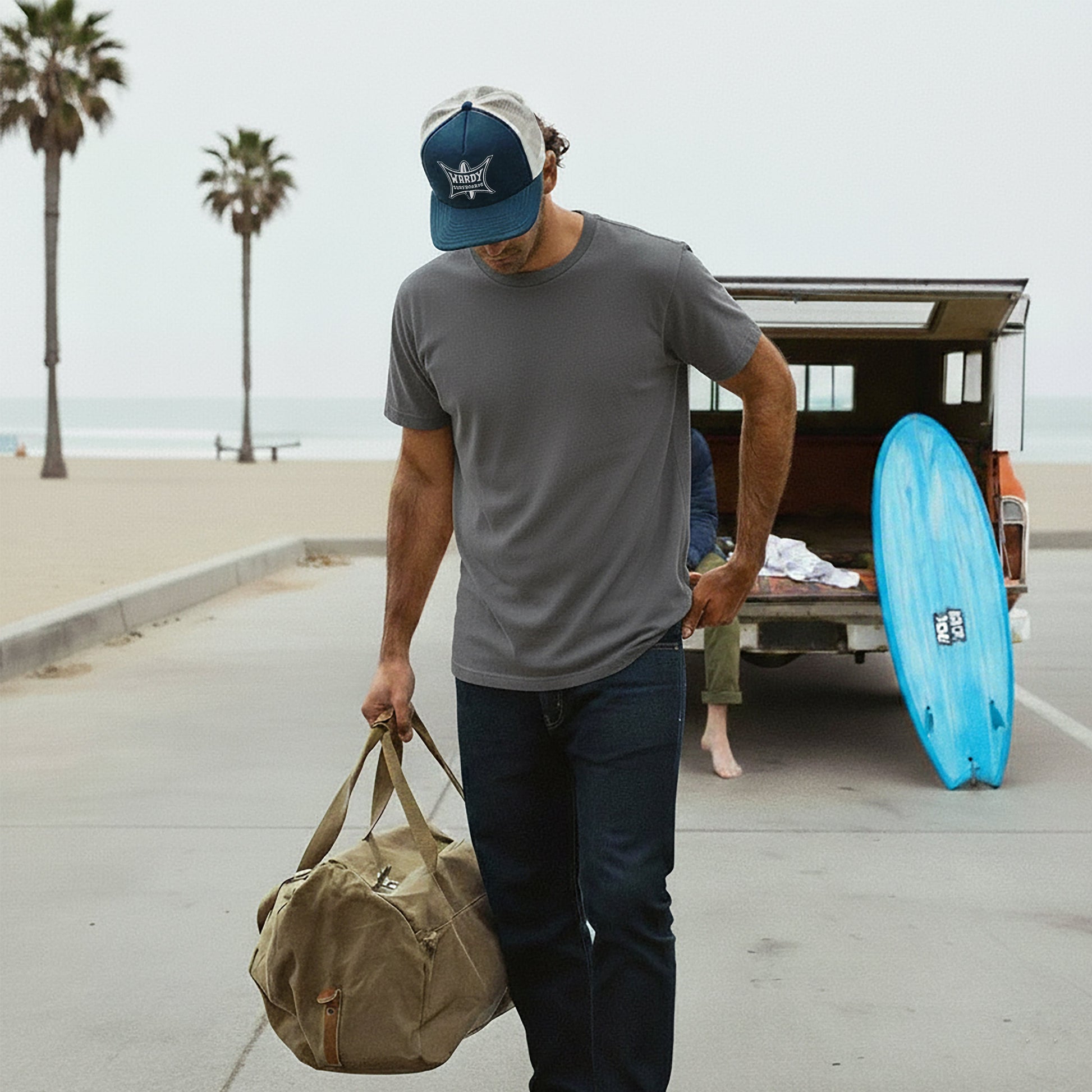 Man walking with a duffel bag on a beachside road with a truck and surfboard in the background. Man wears Navy/white WardySurfboard Trucker hat.