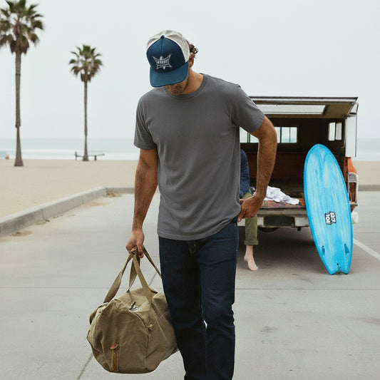 Man walking with a duffel bag on a beachside road with a truck and surfboard in the background. Man wears Navy/white WardySurfboard Trucker hat.